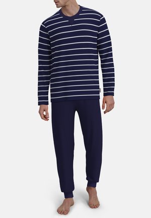 Mann trägt navyblaues, langärmeliges gestreiftes Oberteil und passende navyblaue Hose, steht barfuß vor schlichter Hintergrund.