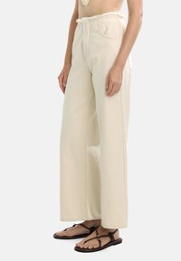 Kremowe spodnie o szerokich nogawkach z postrzępioną talią, wykonane z denimu. Posiadają wysoki stan i prosty krój. W zestawie z brązowymi płaskimi sandałami.