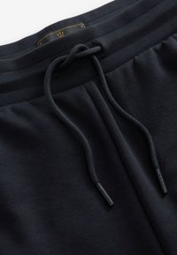 Pantalons de survêtement noirs en tissu doux et extensible. Ils comportent une taille élastique avec cordon de serrage et une étiquette dorée sur la taille.