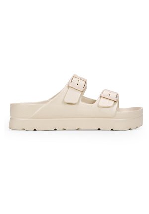 Sandalia deslizante beige con dos correas ajustables, parte superior texturizada y una suela gruesa, moldeada, con un patrón de ondulaciones.