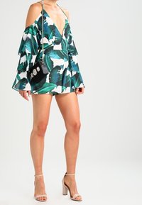 Květinový overal s odkrytými rameny v zeleném a bílém listovém potisku. Černá kabelka s dekoračními detaily. Nahé sandály na blokovém podpatku.
