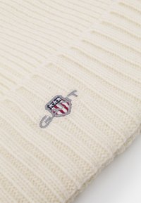 Cream-colored knit beanie with ribbed texture. Features an embroidered patch with a shield design in red, white, and blue accents.