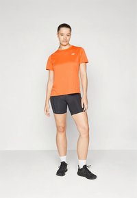 Oransje atletisk t-skjorte og svarte shorts, begge tettsittende. T-skjorten har en reflekterende logo; shortsen har glatt tekstur og går til kneet.