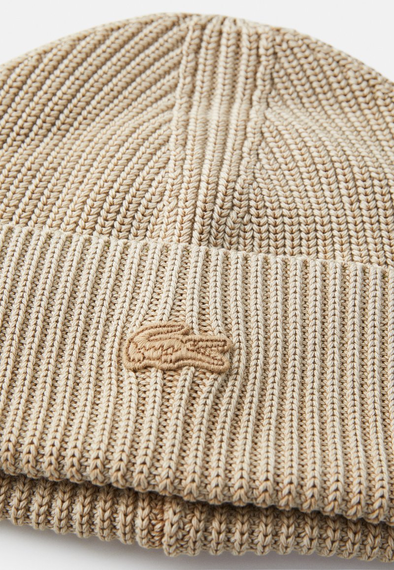Berretto lavorato a maglia in beige con texture a coste e risvolto. Presenta un piccolo logo ricamato in un colore beige abbinato.