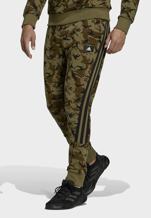 Homme portant un pantalon de survêtement Adidas camouflage vert olive et marron avec des bandes noires et des baskets Adidas noires, debout sur un fond blanc.