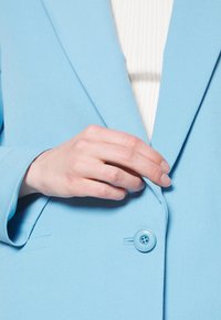 Blazer azzurro chiaro con tessuto testurizzato, bavero a punta e chiusura con un singolo bottone blu. Mano posizionata sul bavero del blazer.