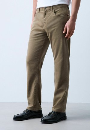 Man draagt beige recht vallende broek, zwarte loafers en een wit overhemd, staand op een effen lichte achtergrond.
