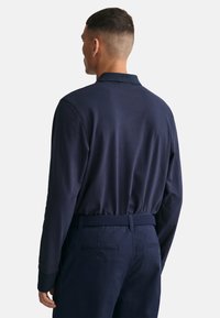 Tengerészkék, hosszú ujjú póló lapos gallérral és texturált mandzsettával, sötét nadrággal, amelyhez szőtt öv tartozik, hátulról nézve.