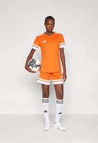 Oransje fotballutstyr med hvite striper på ermene og shortsen; kombinert med hvite sokker med svarte striper og en svart-hvitt mønstrete ball.