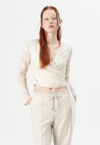 Blonde spiss bluse i off-white med lange blonde ermer, paret med beige bukser med snøring og elastisk liv. 