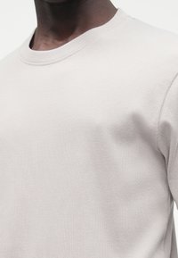 Cămașă gri deschis cu mâneci lungi, cu dungi, cu decolteu rotund, având o textură moale și linii orizontale subtile. Vedere de aproape a țesăturii.