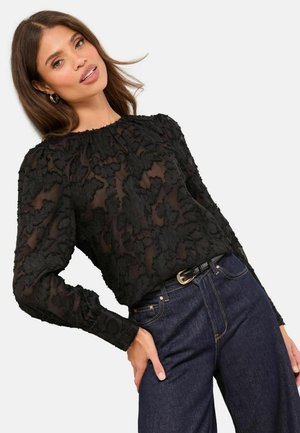 Fekete blúz texturált virágos csipkemintával, hosszú ujjakkal és fodros nyakkivágással. Magas derekú sötét farmernadrággal párosítva.