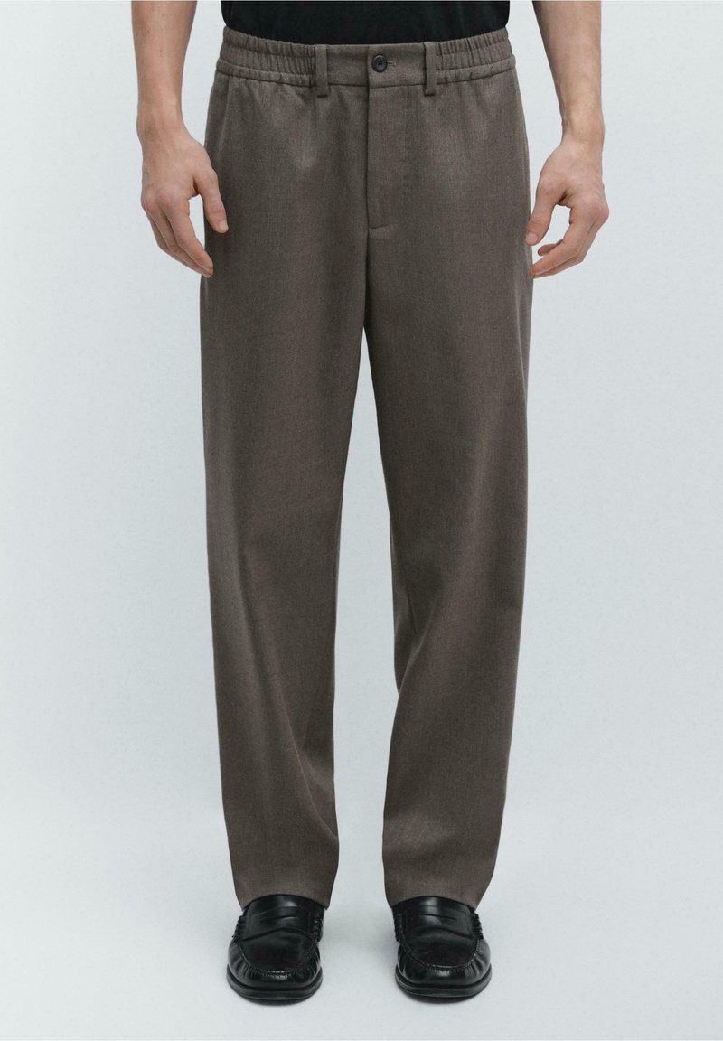 Homme portant un pantalon marron coupe décontractée avec ceinture élastique et mocassins en cuir noirs, debout devant un fond clair uni.