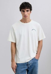 Camiseta de algodón blanca con cuello redondo y mangas cortas, con texto bordado en azul en la parte izquierda del pecho. Usada con jeans azules.
