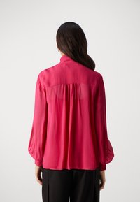 Fuchsia bluse med lange, puffede ærmer og høj krave. Gennemsigtig panel på den øvre ryg med indsamlet detalje, der tilføjer tekstur til designet.