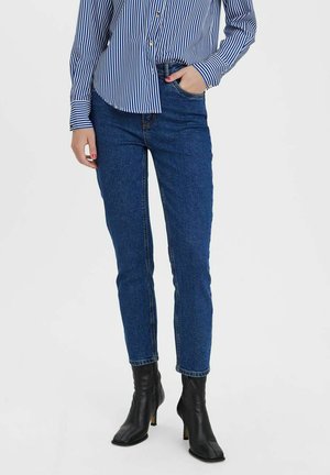Jeans in denim blu con vestibilità slim, con vita media e cinque tasche, abbinati a stivali neri con tacco e una camicia a righe blu.