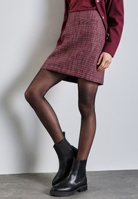 Minifalda de tweed burdeos con un patrón texturizado, que presenta un detalle de botones en el lateral. Combinada con botas negras hasta el tobillo y medias negras transparentes.