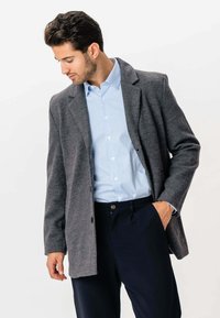 Mand iført en grå blazer, en lyseblå stribet skjorte og mørke bukser, står med den ene hånd i lommen og ser ned til venstre.