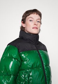 Casaco puffy verde e preto com gola alta, textura brilhante, fecho de zip e um bloqueio de cores distinto, apresentando um leve brilho.