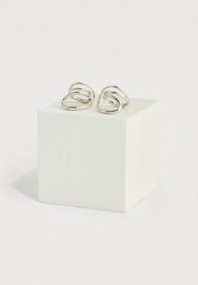 Ζευγάρι ασημένια σκουλαρίκια σε σχήμα σπείρας, τοποθετημένα πάνω σε άσπρο κύβο με φόντο ένα απλό λευκό φόντο.