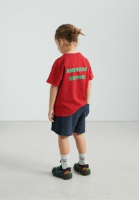Rood katoenen T-shirt met groene tekst "AMERICAN VINTAGE" op de achterkant, gecombineerd met donkerblauwe gestructureerde shorts en zwarte sportschoenen.