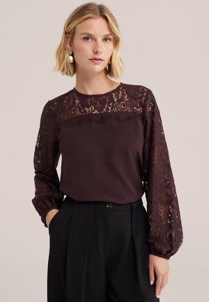 Femme blonde portant un chemisier en dentelle violet foncé à manches longues et un pantalon noir taille haute, debout devant un fond clair.