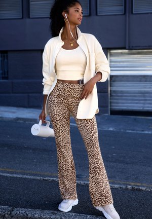 Cream zip-up jacket, beige crop top, brown leopard print flared pants, and white sneakers. Holding a white water bottle.