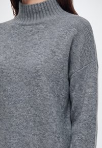 Gros plan d'une personne portant un pull à col roulé gris côtelé en tissu tricoté doux et texturé.