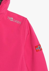 Rožinė striukė su minkšta, lygi texture. Yra "TROLLKIDS" logotipas ir Norvegijos vėliavos užsegtukas ant rankovės. Siūlės sukurtos mašinos.