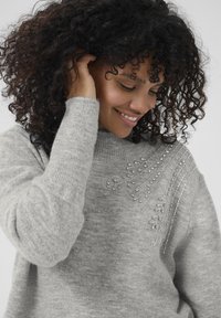 Pelēks džemperis, izgatavots no mīksta auduma, ar augstu apkakli un dekoratīvām pērlītēm un līniju rotājumiem uz vienas pleca.