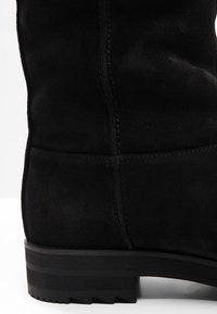 Černé semišové vyšší boty po kolena s texturovaným povrchem, kontrastním prošíváním a pevným, drsným černým gumovým podpatkem.