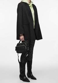 Osoba oblečená v černém saku, svetru světle zelené barvy, černých legínách s leopardím potiskem, černých vysokých botách ke kolenům a držící malou černou tašku.