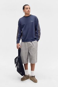 Tmavě modrá mikina s šedými detaily, kárované kraťasy v bílé a šedé barvě, béžové boty a černá taška přes rameno s bílým logem.