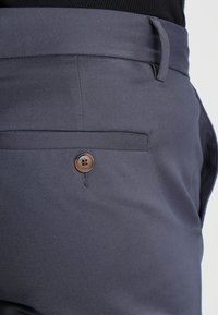 Dark gray trousers feature a smooth texture, a single back pocket with a brown button closure, and belt loops on the waistband.
