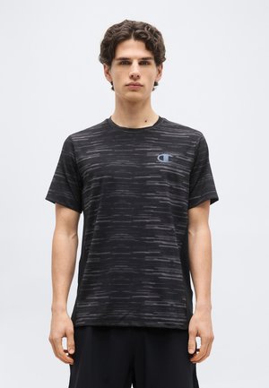 Νεαρός άνδρας που φοράει αθλητική κοντομάνικη μπλούζα σε μαύρο και γκρι με μοτίβο και μικρό μπλε λογότυπο στο αριστερό στήθος, στέκεται μπροστά σε ένα απλό φόντο.