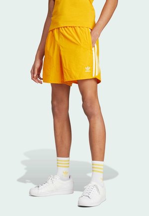 Osoba ubrana w jaskrawożółte spodenki Adidas z białymi paskami, białe skarpetki Adidas z żółtymi paskami i białe trampki, stojąca.