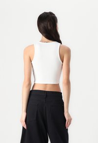 Bela pritrjena majica brez rokavov z rebrasto teksturo, zaobljenim izrezom in debelimi naramnicami, kombinirana z visokim pasom črnih hlač.