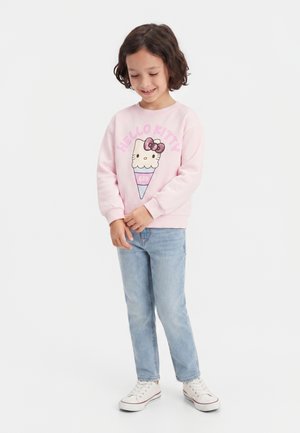 Enfant souriant portant un sweat rose Hello Kitty, un jean bleu clair et des baskets blanches, debout les mains jointes devant un fond uni.