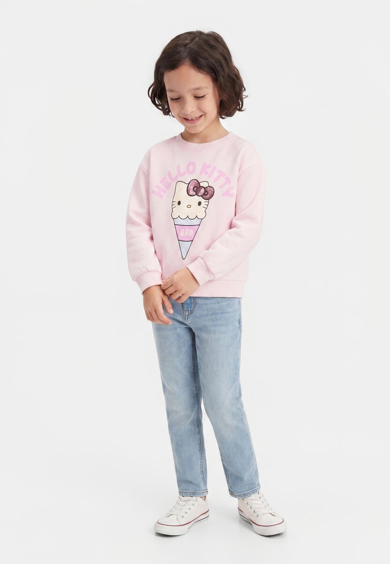 Enfant souriant portant un sweat rose Hello Kitty, un jean bleu clair et des baskets blanches, debout les mains jointes devant un fond uni.