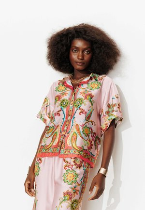 Femme à la peau foncée aux cheveux bouclés et volumineux porte une chemise et un pantalon en soie rose assortis avec des motifs floraux colorés et cachemire, des bijoux en or.