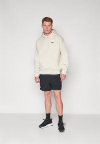 Lichtbeige hoodie met een buidelzak en een trekkoordcapuchon, gecombineerd met zwarte shorts en zwarte sportschoenen. Gladde textuur, casual ontwerp.