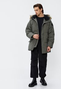 Olivgrüne Pufferjacke mit fellgefütterter Kapuze, vorderem Reißverschluss und Seitentaschen. Getragen über einem schwarzen Shirt und kombiniert mit schwarzen Cargo-Hosen.