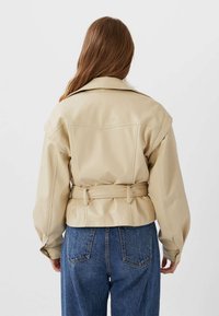 Beige leren jas met een brede kraag, een ceintuur op de taille en volumineuze mouwen, gecombineerd met hoog-wijd blote blauwe denim jeans.