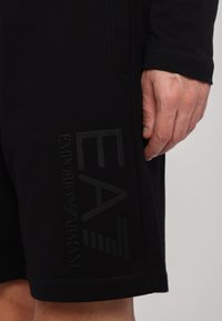 Pantalones cortos negros de tejido suave con un logo goma "EA" y "EMPORIO ARMANI" en el lateral, con un diseño aerodinámico y detalles de costura.