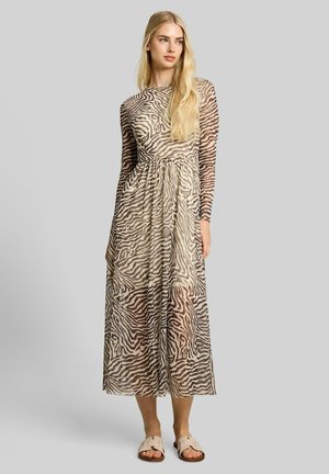 Vrouw met lang blond haar draagt een doorschijnende, lange mouw midi-jurk met zebraprint en beige instapsandalen tegen een effen grijze achtergrond.