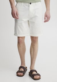 Shorts bianchi di cotone con orli arrotolati, dotati di chiusura a bottone e tasche frontali. Indossati con sandali marroni, che mostrano superfici morbide e strutturate.