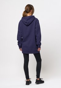 Femme debout, dos tourné, portant un sweat à capuche bleu foncé oversize, des leggings noirs et des chaussures de sport noires sur un fond uni.