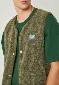 Green fleece vest with a textured surface, snap buttons, contrasting olive trim, and a small blue and white branded patch. Layered over a green shirt.