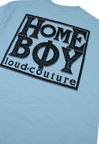 Šviesiai mėlynas marškinėliai su dideliu juodu grafiku, kuriame yra tekstas "HOME BOY loud.couture" paryškintomis blokinėmis raidėmis ir unikaliu apipavidalinimu.