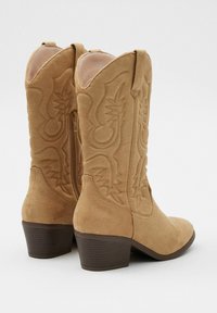 Botas de tobillo de ante color topo con un diseño cosido, punta redondeada y un tacón bajo. Cuentan con cremallera lateral y tiradores para ponérselas fácilmente.